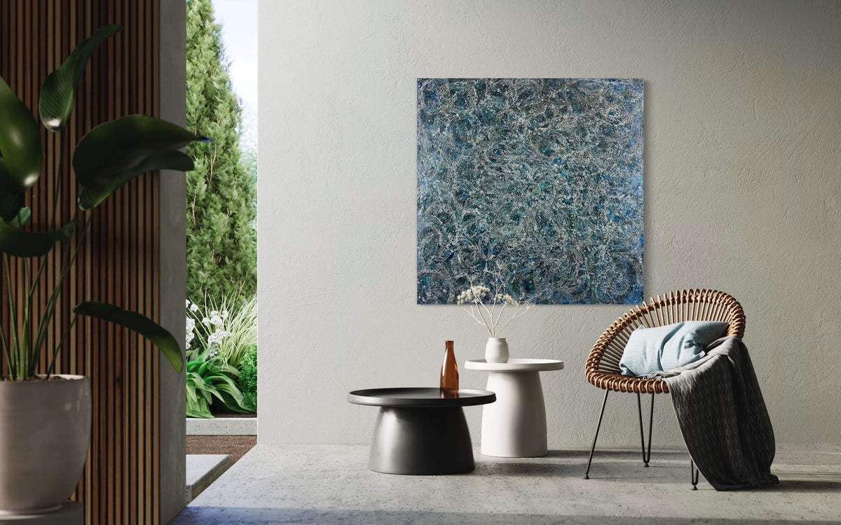 Original abstract encaustic painting in deep blues and white tones with lots of visual texture and many lace-like swirls. Displayed on a painted concrete wall above a rattan chair and two side tables in a modern interior