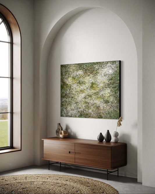 Original abstract encaustic painting with delicate layers of soft white, mossy green and earthy tones. The surface is textured with fine, mist like dots, and organic forms, evoking a sense of gentle movement, growth, and natural stillness displayed in modern interior above a credenza.