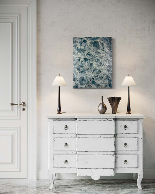 Original abstract encaustic painting in moody blues, whites and it has flow and movement to it to represent the movement of water displayed on a plaster wall above a dresser, flanked by two lamps